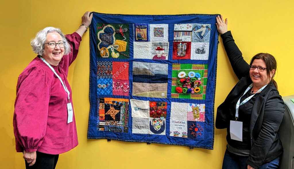 A quilt being held up against a bright plain wall. The person on the left is a female in a bright pink plain top and grey hair and the person on the right is a female with a dark jacket and dark hair. Both are smiling.