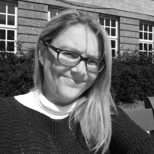 Black and white photo of a white mid aged female with glasses and pale hair, sitting in a green area