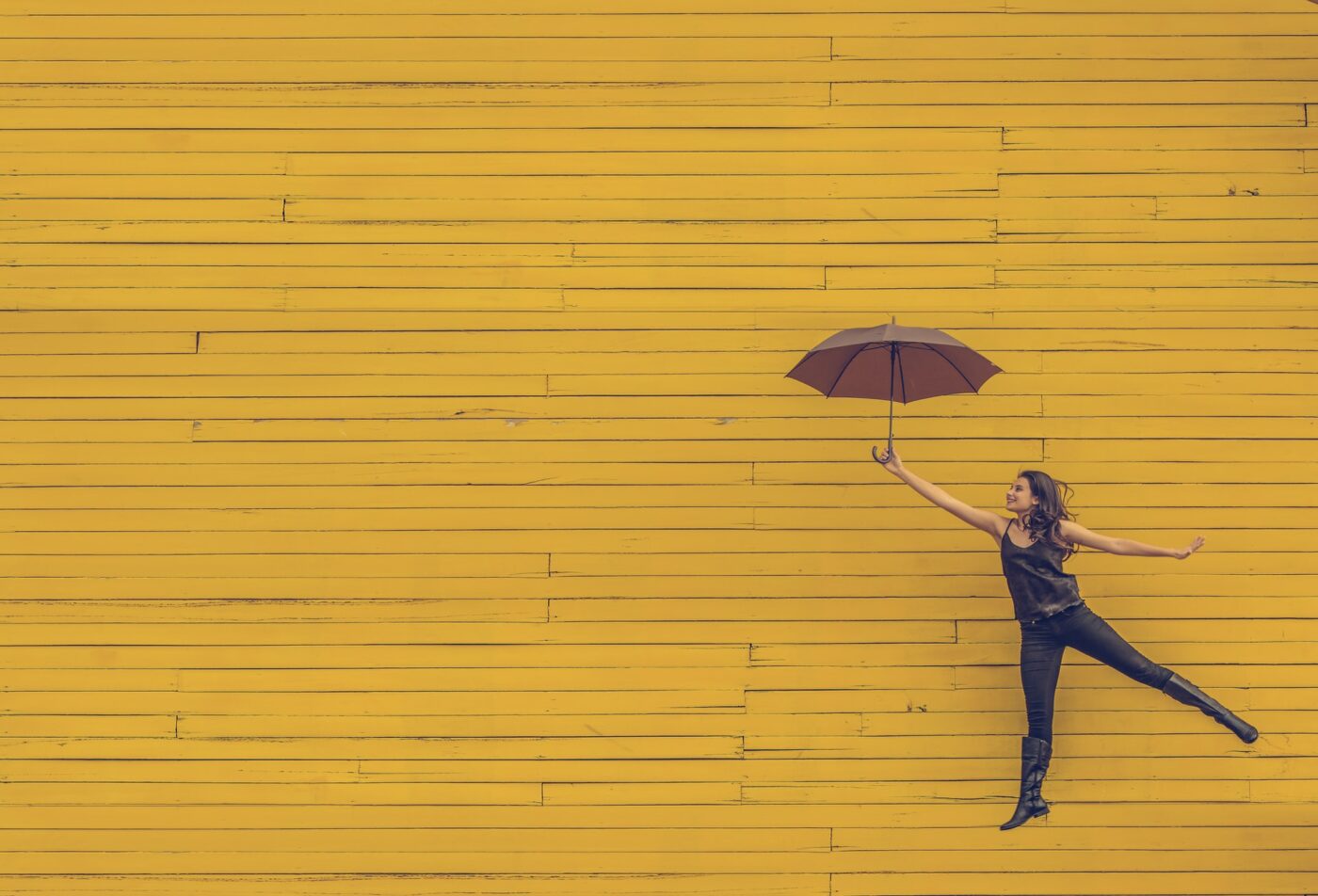 Lady hovering in air with open umbrella against a yellow brick wall